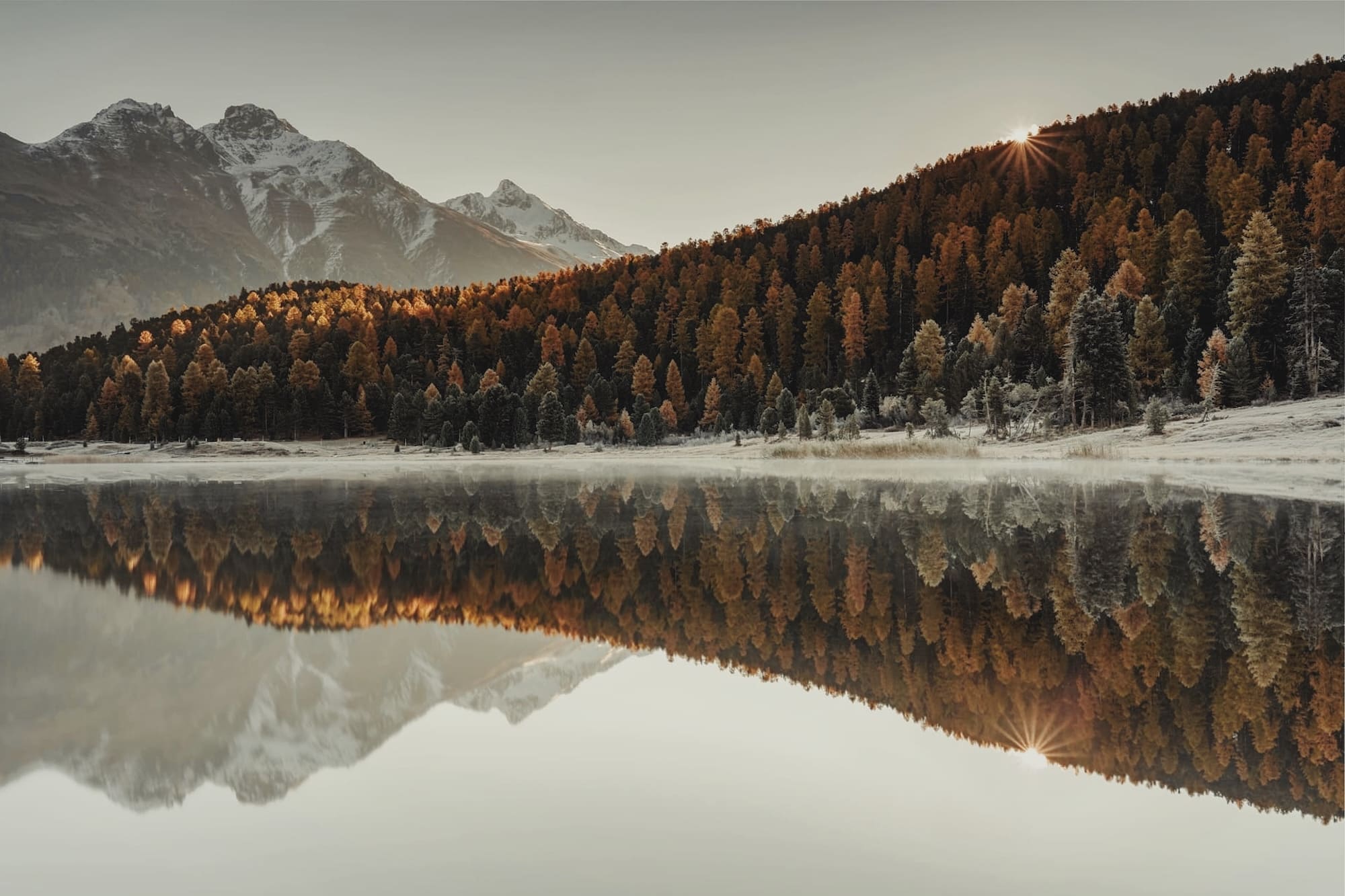 Lac de munte cu reflecție perfectă — pădure de toamnă și vârfuri înzăpezite oglindite în apă liniștită.
